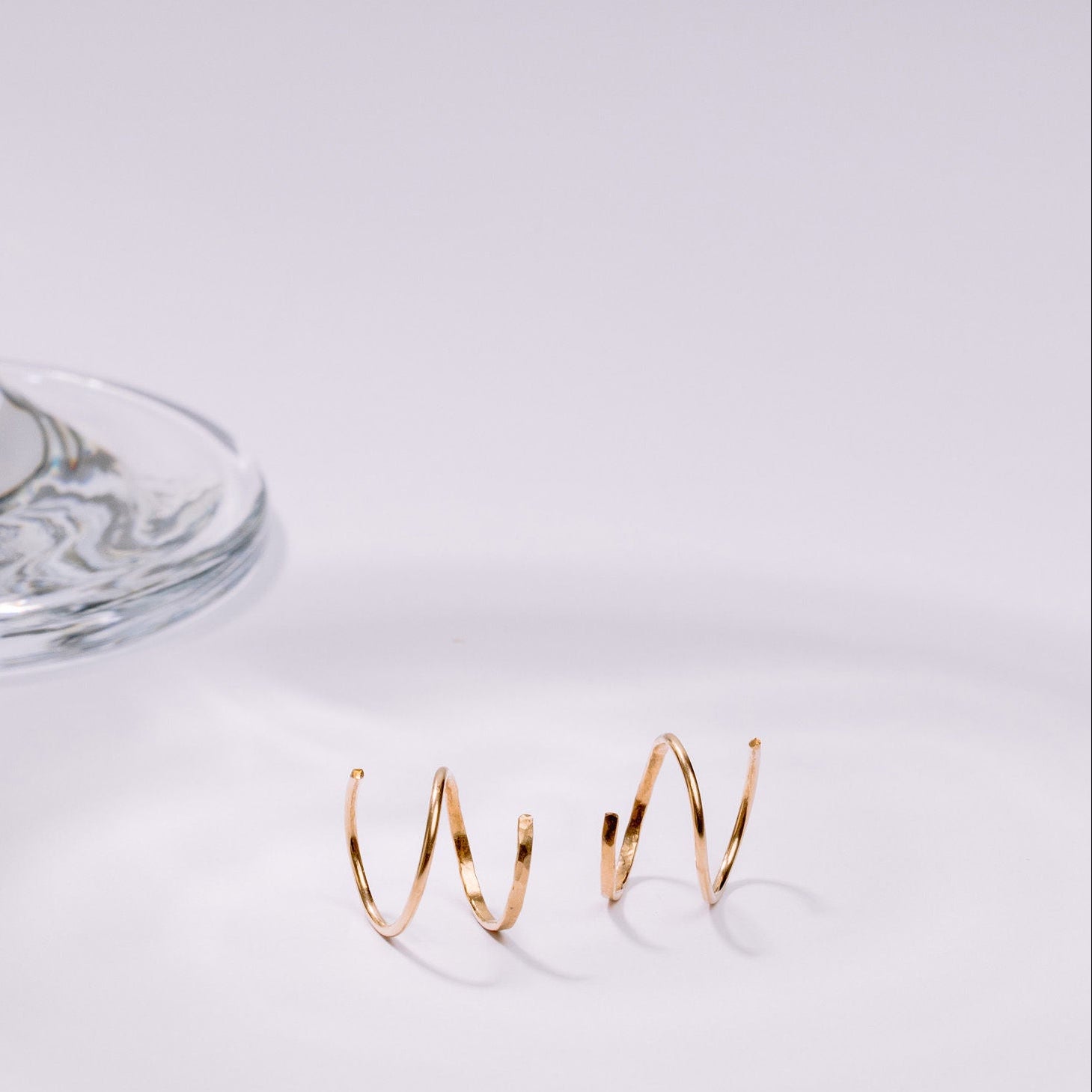 Gold double huggies on a white surface with a glass object in the background