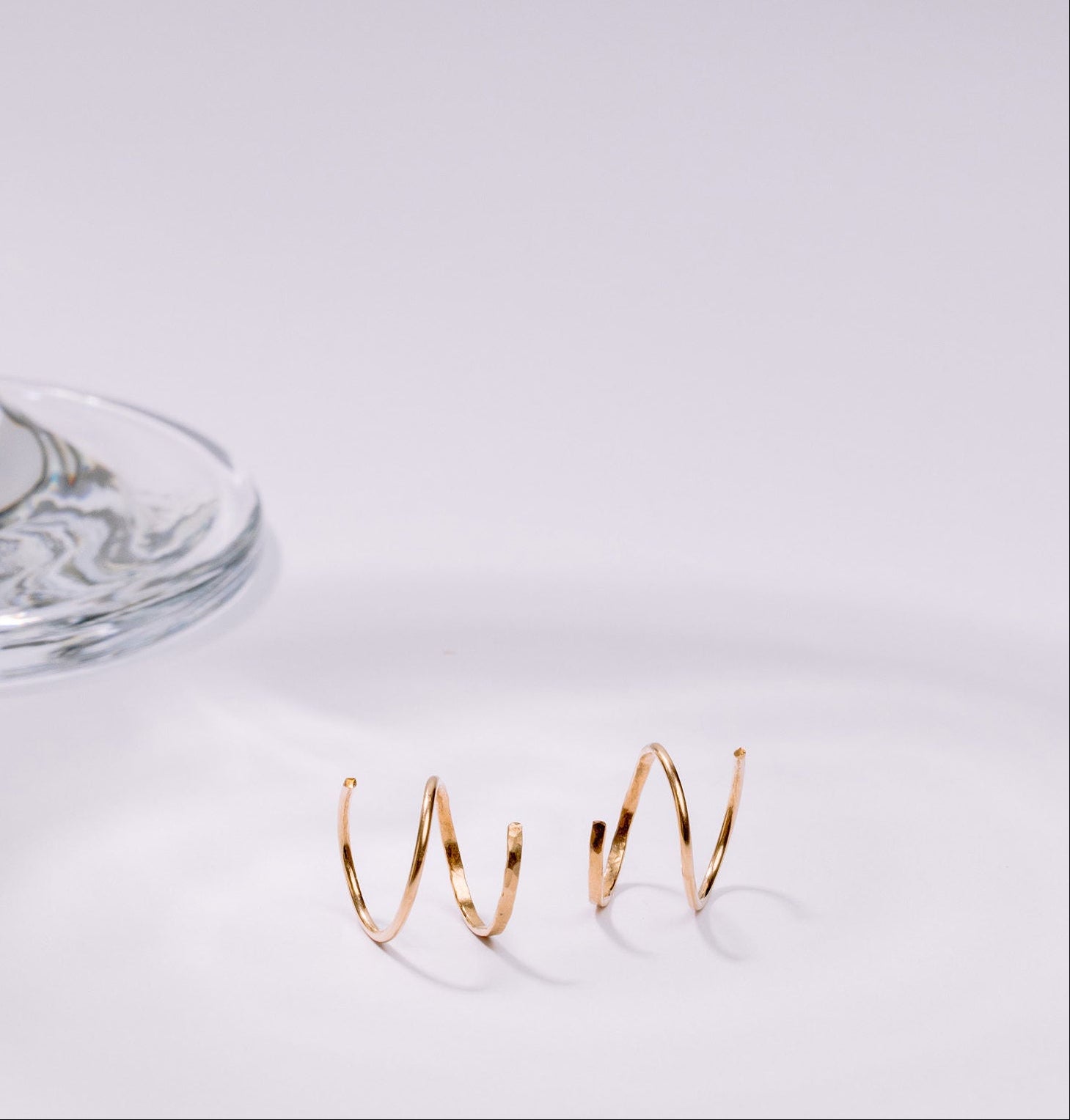 Gold double huggies on a white surface with a glass object in the background