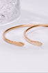 Gold open bangle on a white surface with a glass object in the background