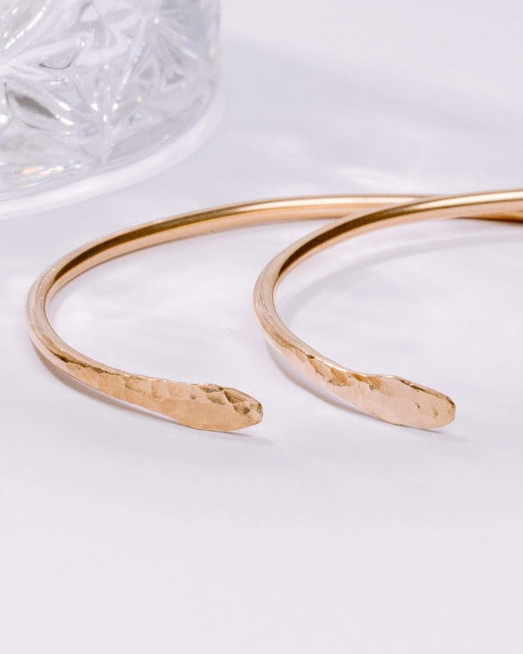 Gold open bangle on a white surface with a glass object in the background