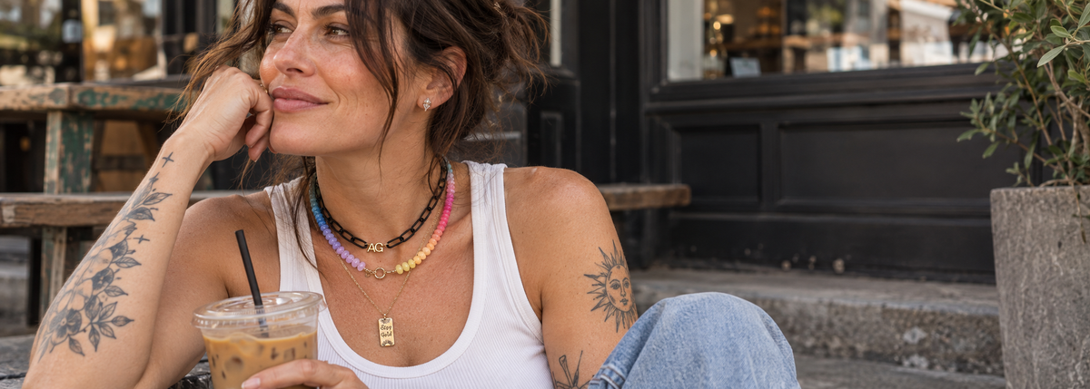 Girl with a white tank on, tattoos, drinking an iced coffee on her front porch steps in the city, with 3 necklaces layered on