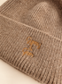 Close-up of a ribbed knit beanie in a warm taupe-beige color with gold embroidered script reading 'Gypsy & Gem' on the cuff.