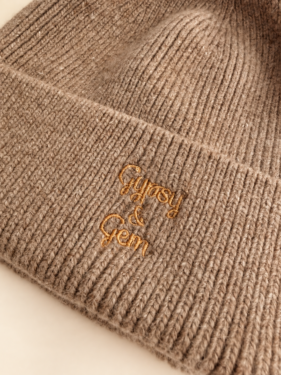 Close-up of a ribbed knit beanie in a warm taupe-beige color with gold embroidered script reading 'Gypsy & Gem' on the cuff.