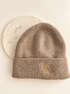 A ribbed knit beanie in taupe-brown with a small gold embroidered logo on the cuff, displayed against a white background with a round marble prop.