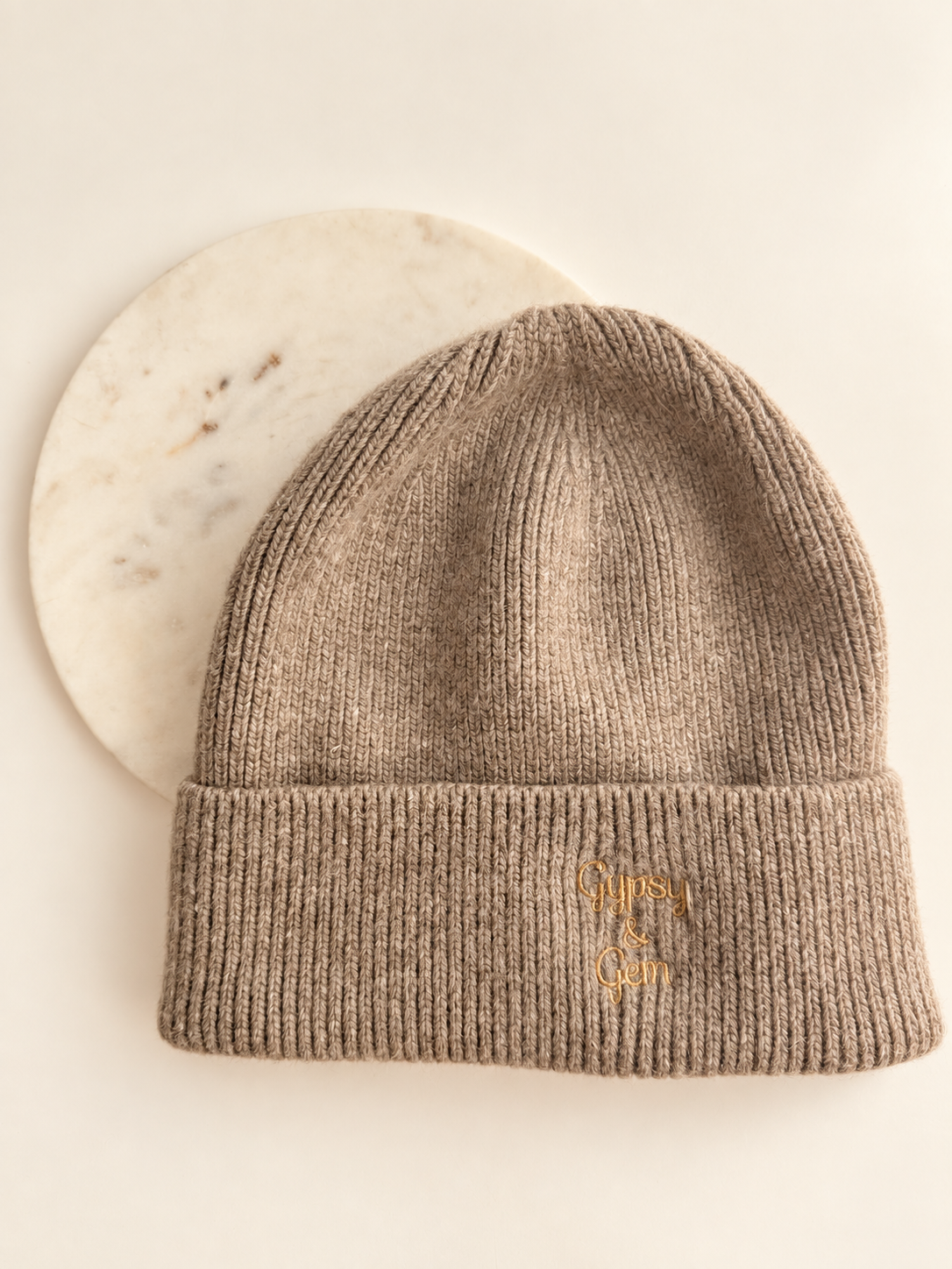 A ribbed knit beanie in taupe-brown with a small gold embroidered logo on the cuff, displayed against a white background with a round marble prop.