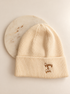 A cream-colored ribbed knit beanie hat with 'Gypsy & Gem' embroidered in brown on the front cuff, displayed on a white background with a round marble coaster behind it.