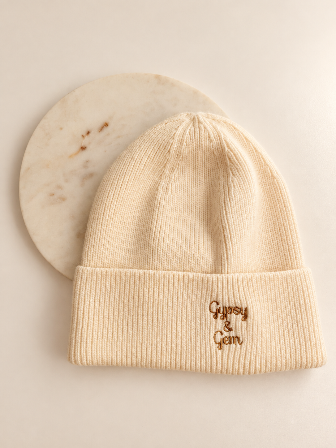 A cream-colored ribbed knit beanie hat with 'Gypsy & Gem' embroidered in brown on the front cuff, displayed on a white background with a round marble coaster behind it.