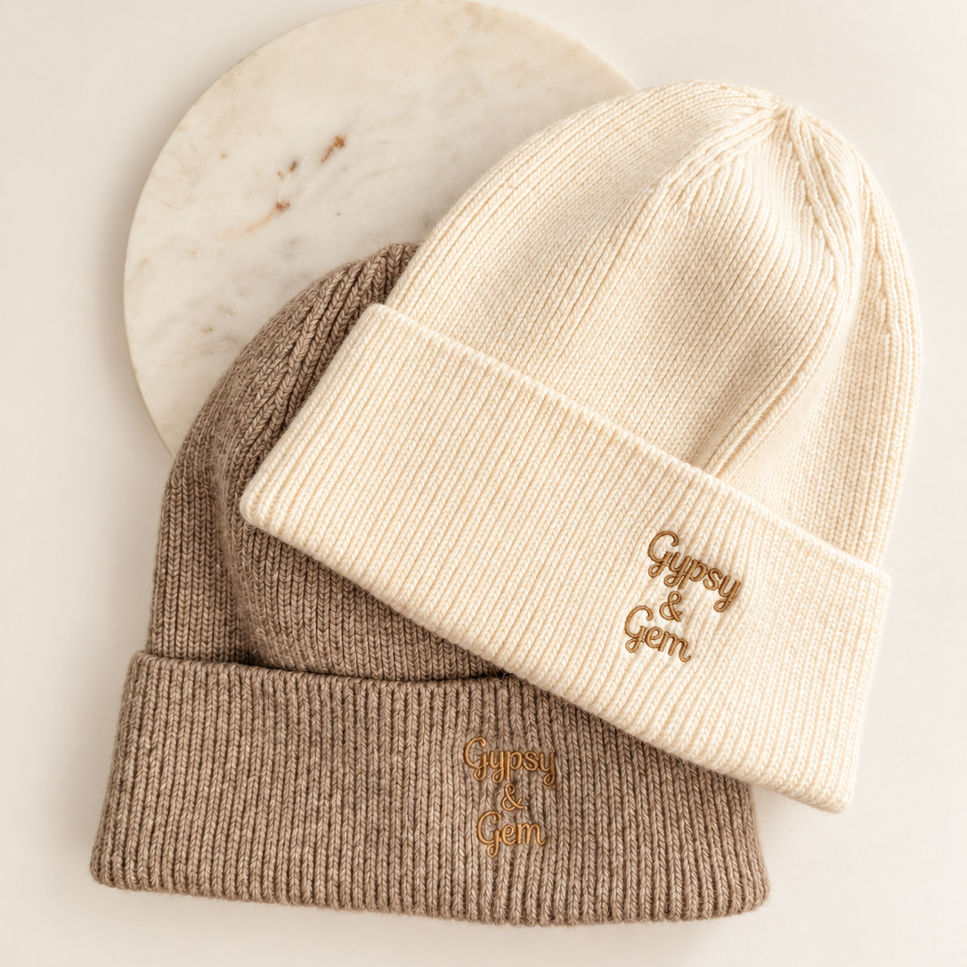 Two ribbed knit beanie hats stacked on a light background, with a cream-colored beanie on top featuring a small gold embroidered logo and a taupe-colored beanie underneath with a matching gold logo. A round white marble coaster is partially visible behind them.