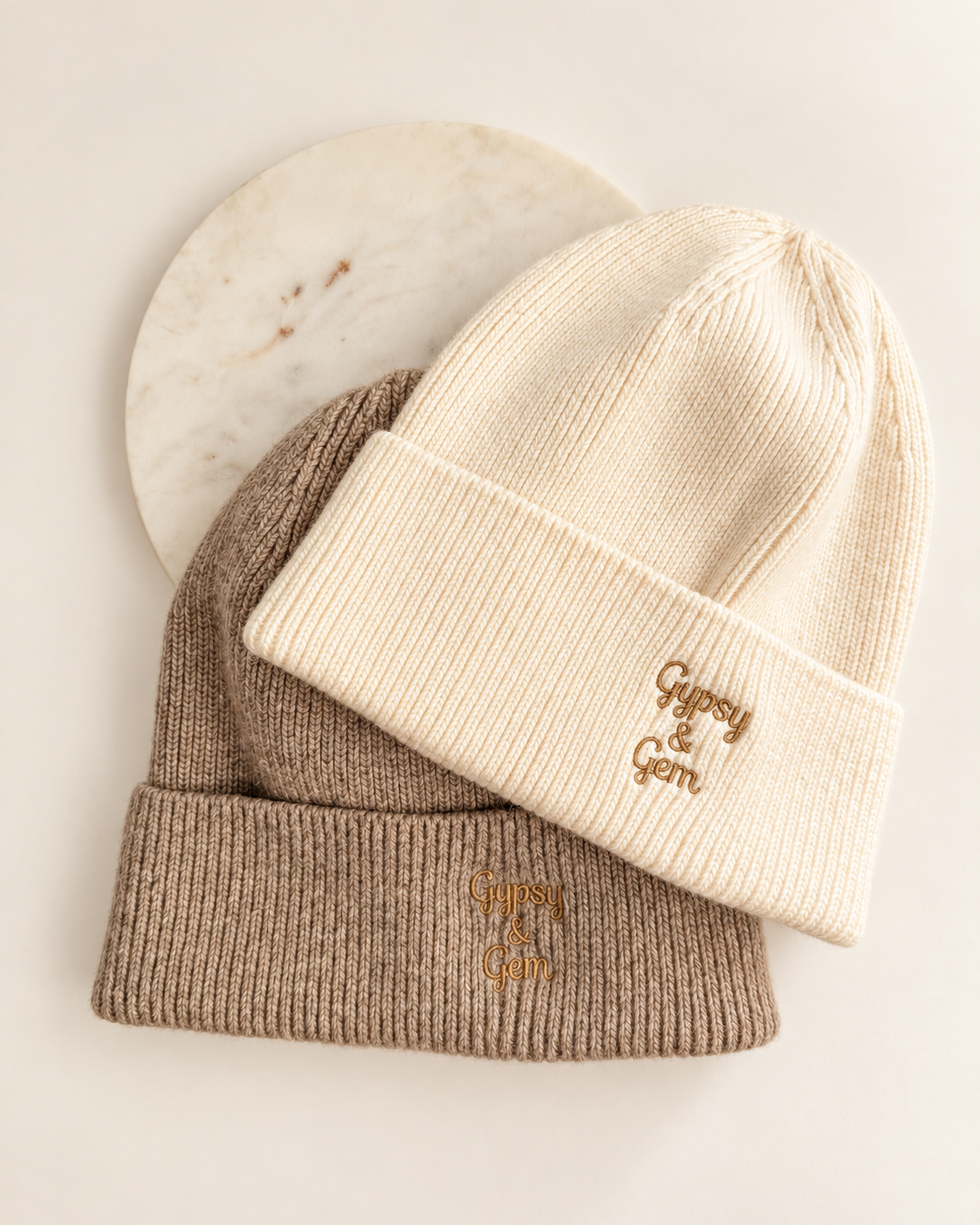 Two ribbed knit beanie hats stacked on a light background, with a cream-colored beanie on top featuring a small gold embroidered logo and a taupe-colored beanie underneath with a matching gold logo. A round white marble coaster is partially visible behind them.