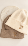 Two ribbed knit beanie hats stacked on a light background, with a cream-colored beanie on top featuring a small gold embroidered logo and a taupe-colored beanie underneath with a matching gold logo. A round white marble coaster is partially visible behind them.