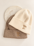 Two ribbed knit beanie hats stacked on a light background, with a cream-colored beanie on top featuring a small gold embroidered logo and a taupe-colored beanie underneath with a matching gold logo. A round white marble coaster is partially visible behind them.