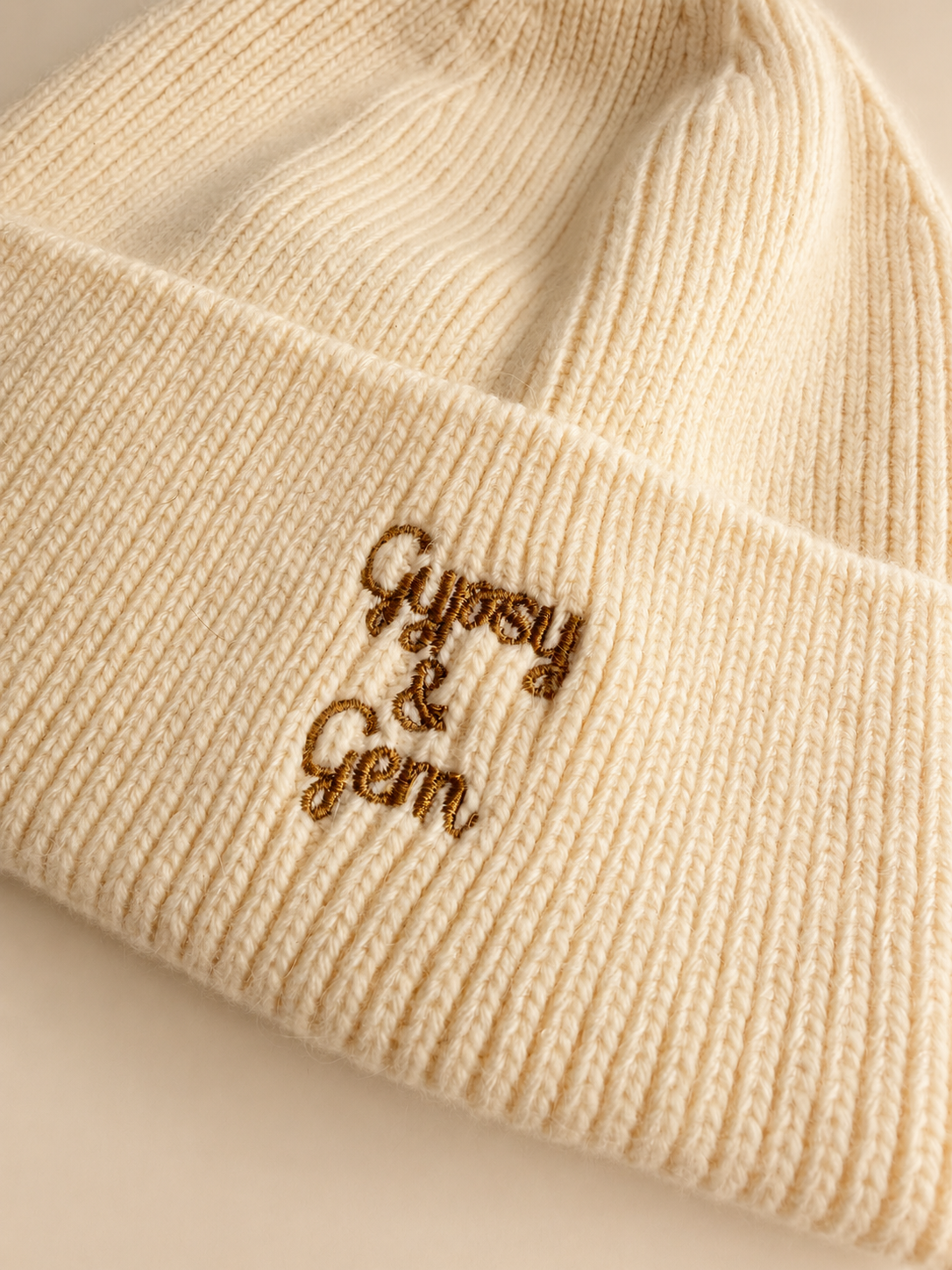 Close-up of a cream-colored ribbed knit beanie with brown embroidered text reading 'Gypsy & Gem' on the cuff.