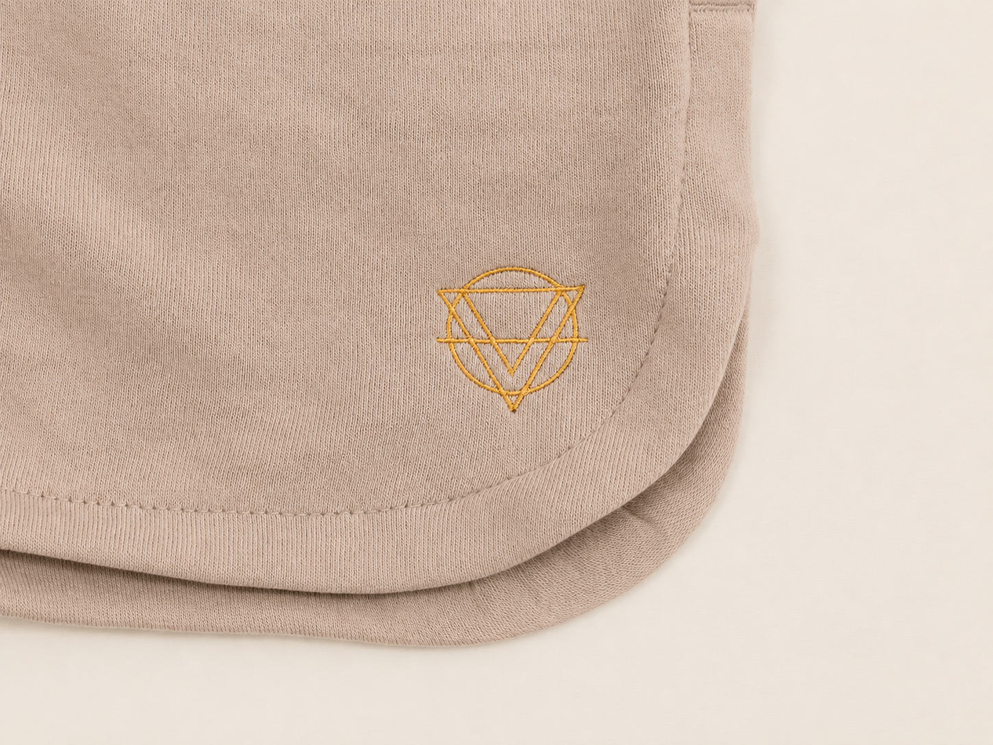 Close-up of a light beige sweatshorts, featuring a small gold embroidered geometric symbol on the lower right.