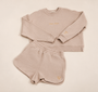 A two-piece women's loungewear set in a light beige or oatmeal color, consisting of a long-sleeve sweatshirt and matching shorts, laid flat on an ivory background