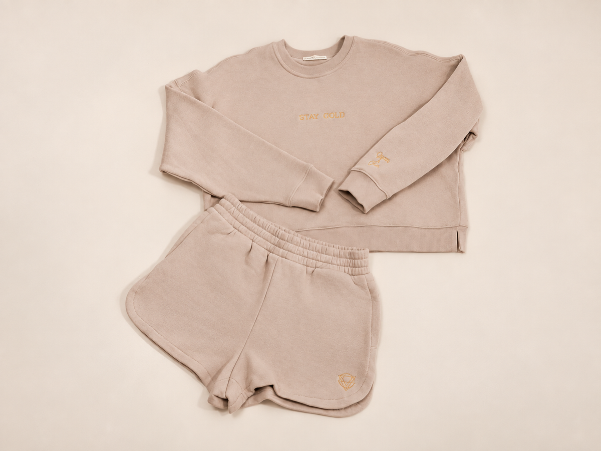A two-piece women's loungewear set in a light beige or oatmeal color, consisting of a long-sleeve sweatshirt and matching shorts, laid flat on an ivory background