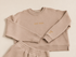A matching two-piece lounge set in a beige color, featuring a crewneck sweatshirt with 'STAY COLD' embroidered on the chest and a small monogram on the cuff, paired with matching sweatshorts on an ivory background.