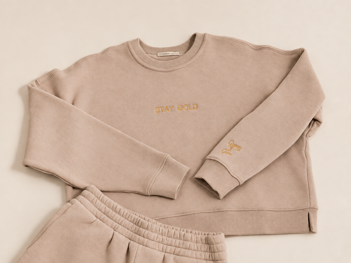 A matching two-piece lounge set in a beige color, featuring a crewneck sweatshirt with 'STAY COLD' embroidered on the chest and a small monogram on the cuff, paired with matching sweatshorts on an ivory background.
