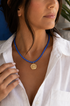 Woman in a white button down blouse wearing a blue stone beaded necklace with a gold round charm with good luck symbols on it. 