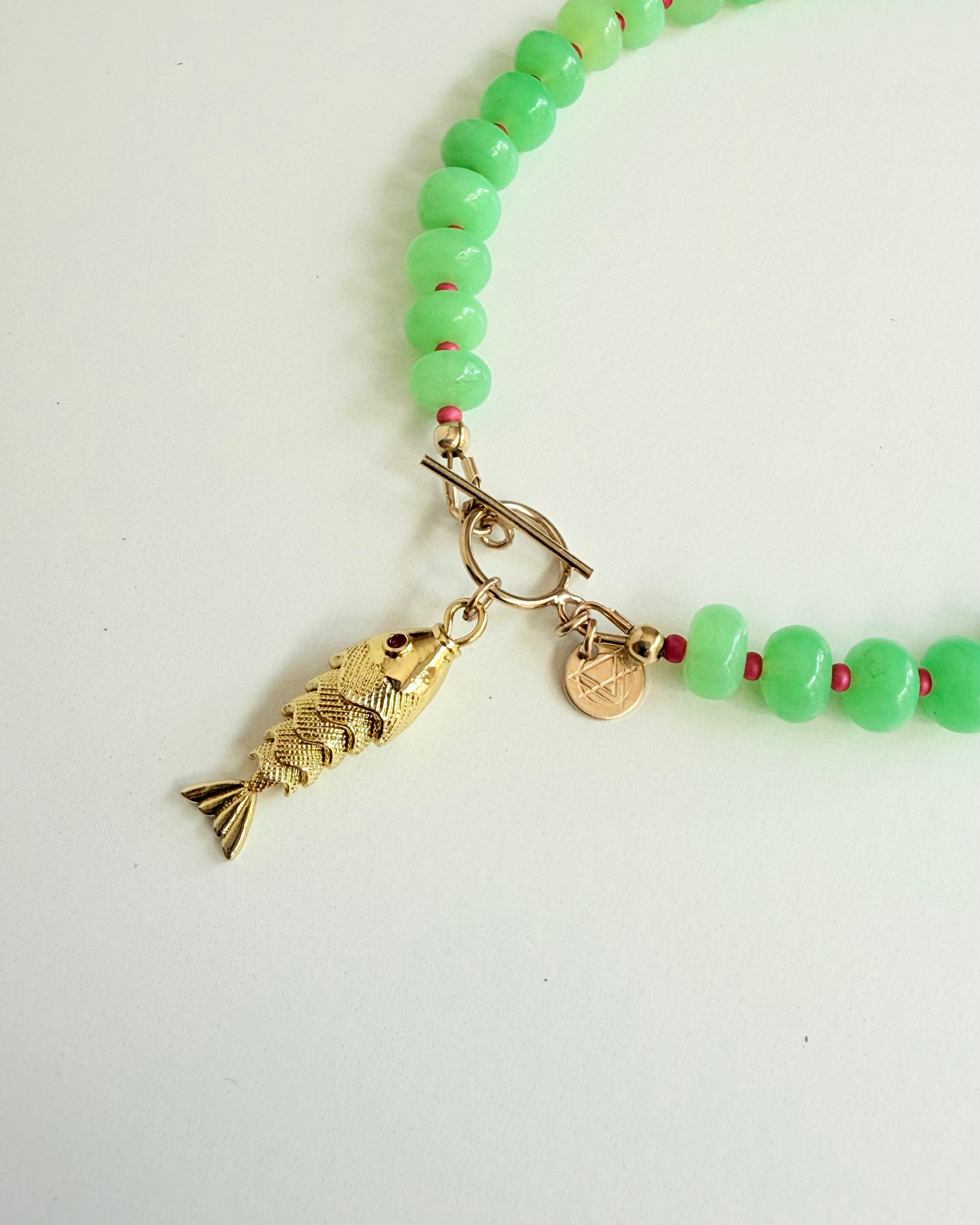 Close up of Green stone necklace with pink seed beads and a gold fish charm with a pink eye on a white background