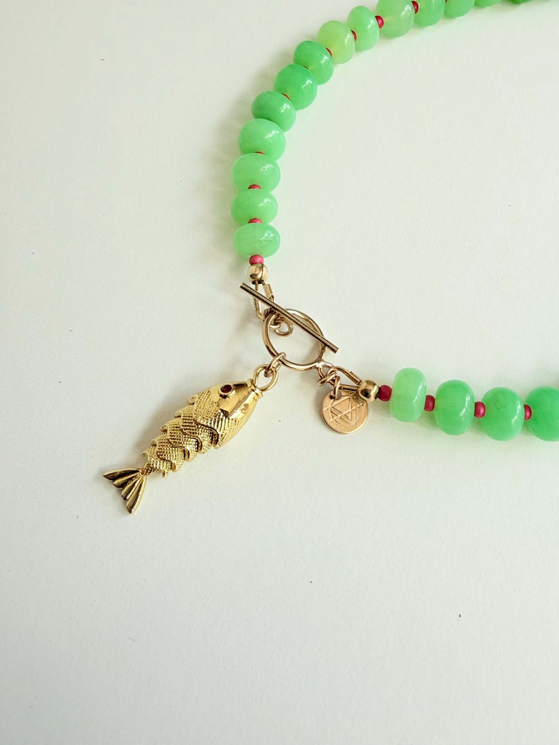 Close up of Green stone necklace with pink seed beads and a gold fish charm with a pink eye on a white background