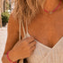 A woman outdoors wearing a pink braided necklace with a gold initial 'AC' pendant and a matching pink bracelet, paired with a cream-colored pleated spaghetti-strap dress.