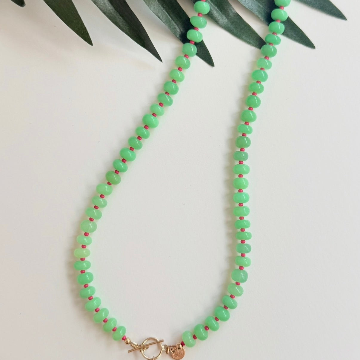 Green stone necklace with pink seed beads and a gold fish charm with a pink eye on a white background with a tropical leaf