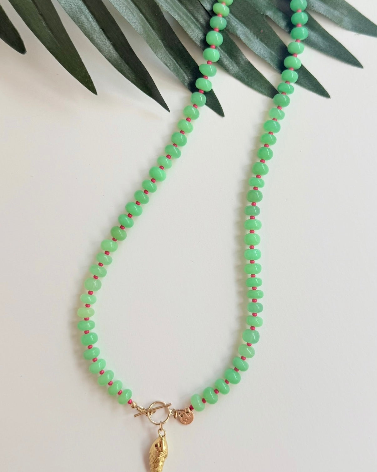 Green stone necklace with pink seed beads and a gold fish charm with a pink eye on a white background with a tropical leaf
