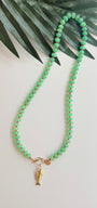 Green stone necklace with pink seed beads and a gold fish charm with a pink eye on a white background with a tropical leaf