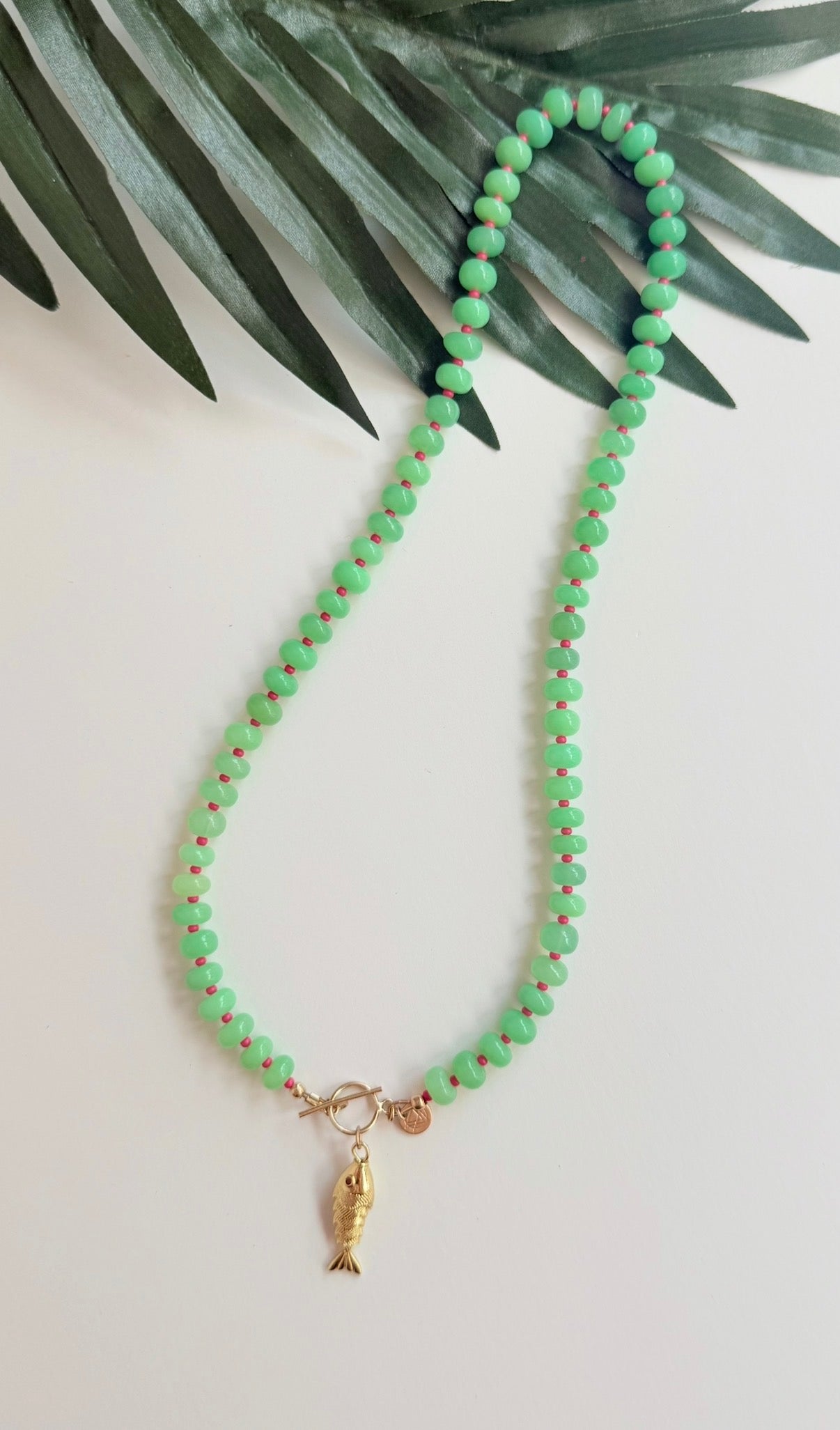 Green stone necklace with pink seed beads and a gold fish charm with a pink eye on a white background with a tropical leaf