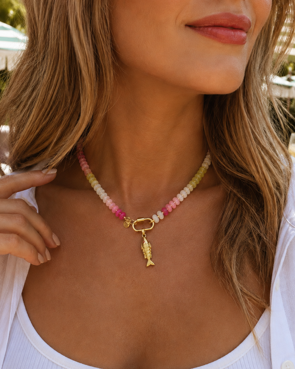 A close-up lifestyle shot of a woman wearing a pastel beaded necklace with a gold clasp and a small gold fish charm, styled with a white ribbed tank top and white button-down shirt.