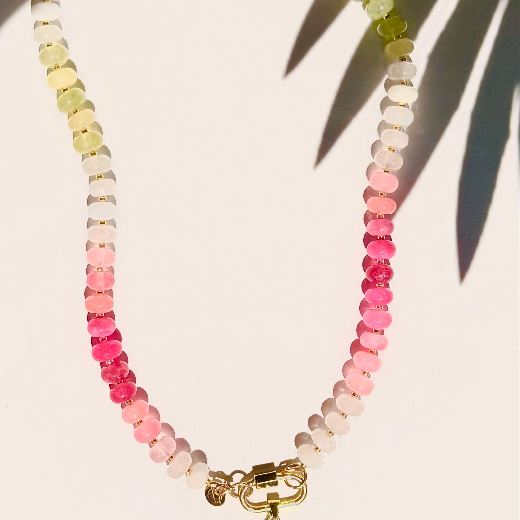 pink and green stone bead necklace with fish charm on white background with tropical leaf shadow