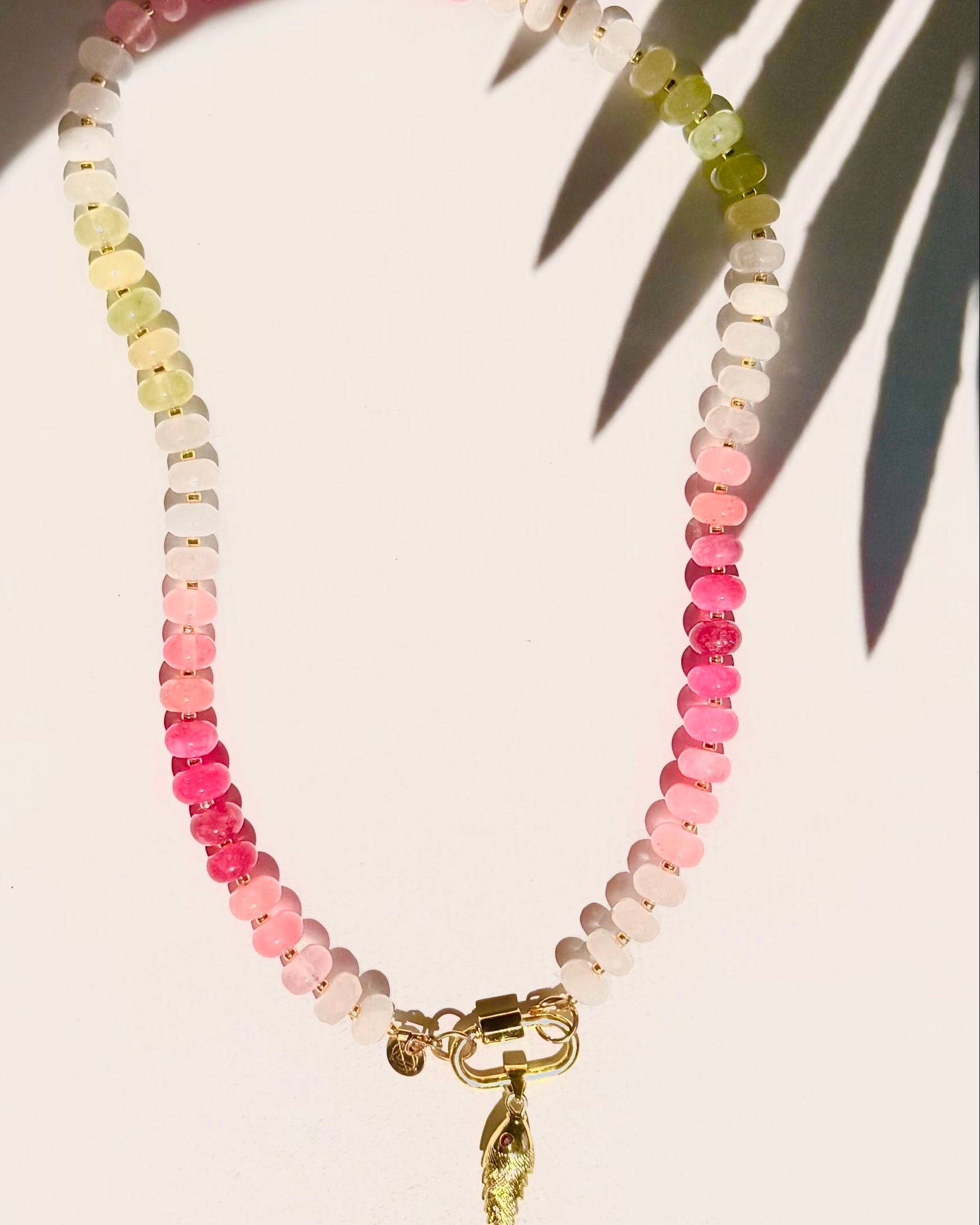 pink and green stone bead necklace with fish charm on white background with tropical leaf shadow