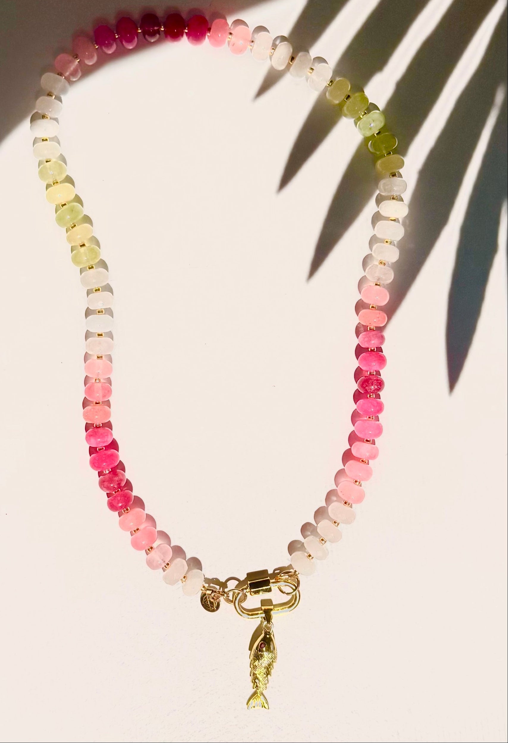 pink and green stone bead necklace with fish charm on white background with tropical leaf shadow