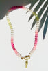 Green pink and white stone beaded necklace with a gold carabiner and gold fish charm on a white background with a tropical leaf shadow