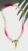 Green pink and white stone beaded necklace with a gold carabiner and gold fish charm on a white background with a tropical leaf shadow