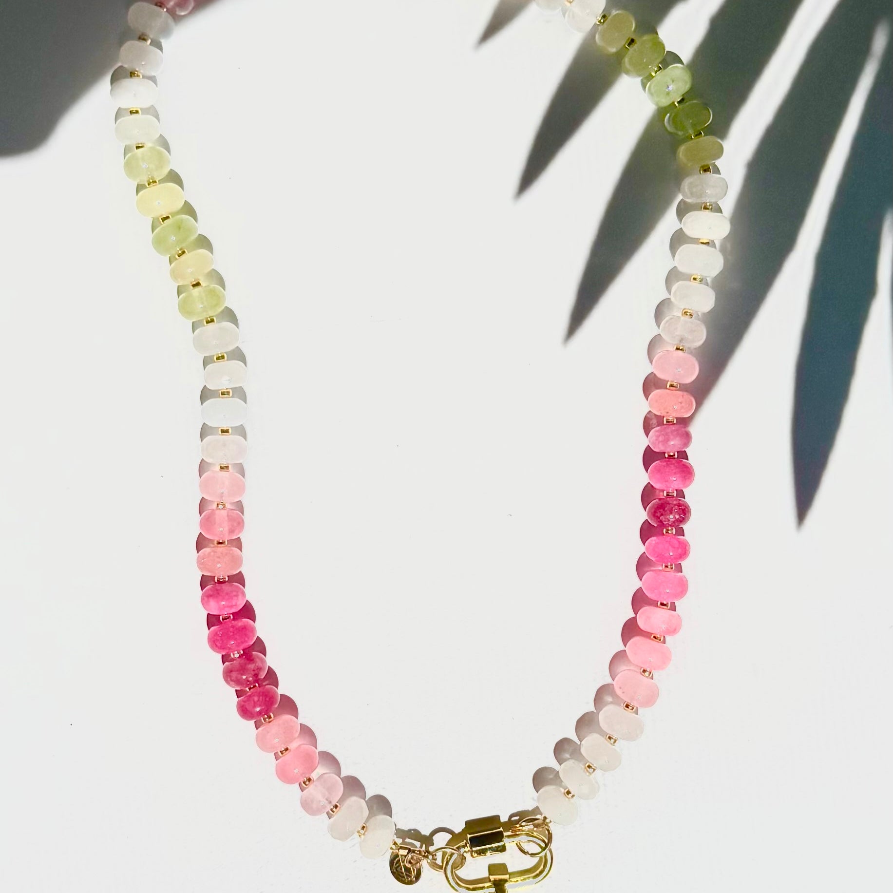 Green pink and white stone beaded necklace with a gold carabiner and gold fish charm on a white background with a tropical leaf shadow