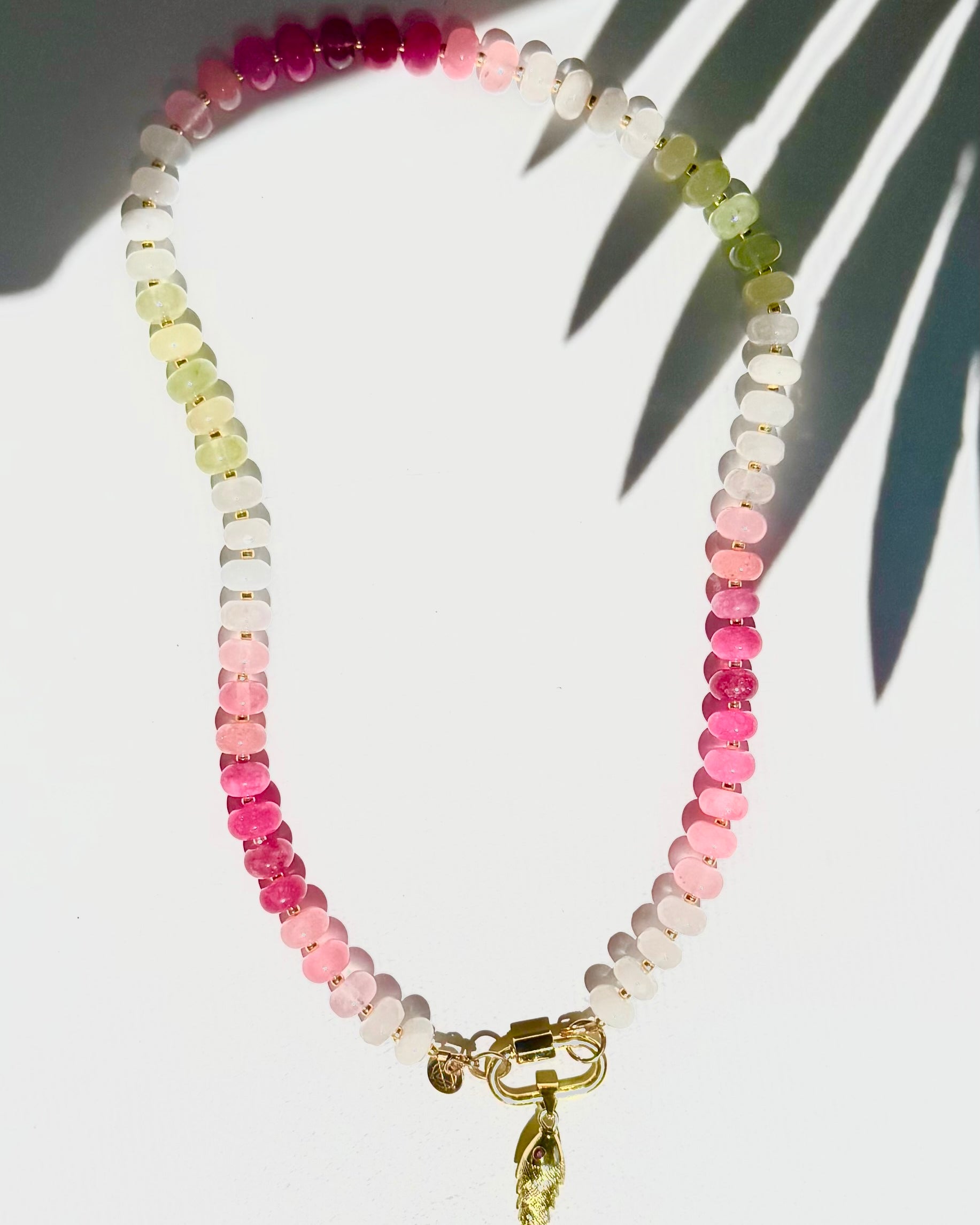 Green pink and white stone beaded necklace with a gold carabiner and gold fish charm on a white background with a tropical leaf shadow