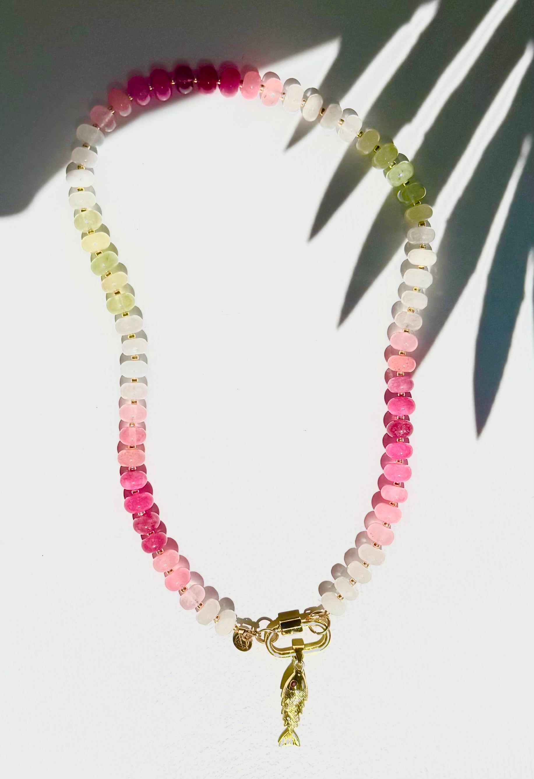 Green pink and white stone beaded necklace with a gold carabiner and gold fish charm on a white background with a tropical leaf shadow