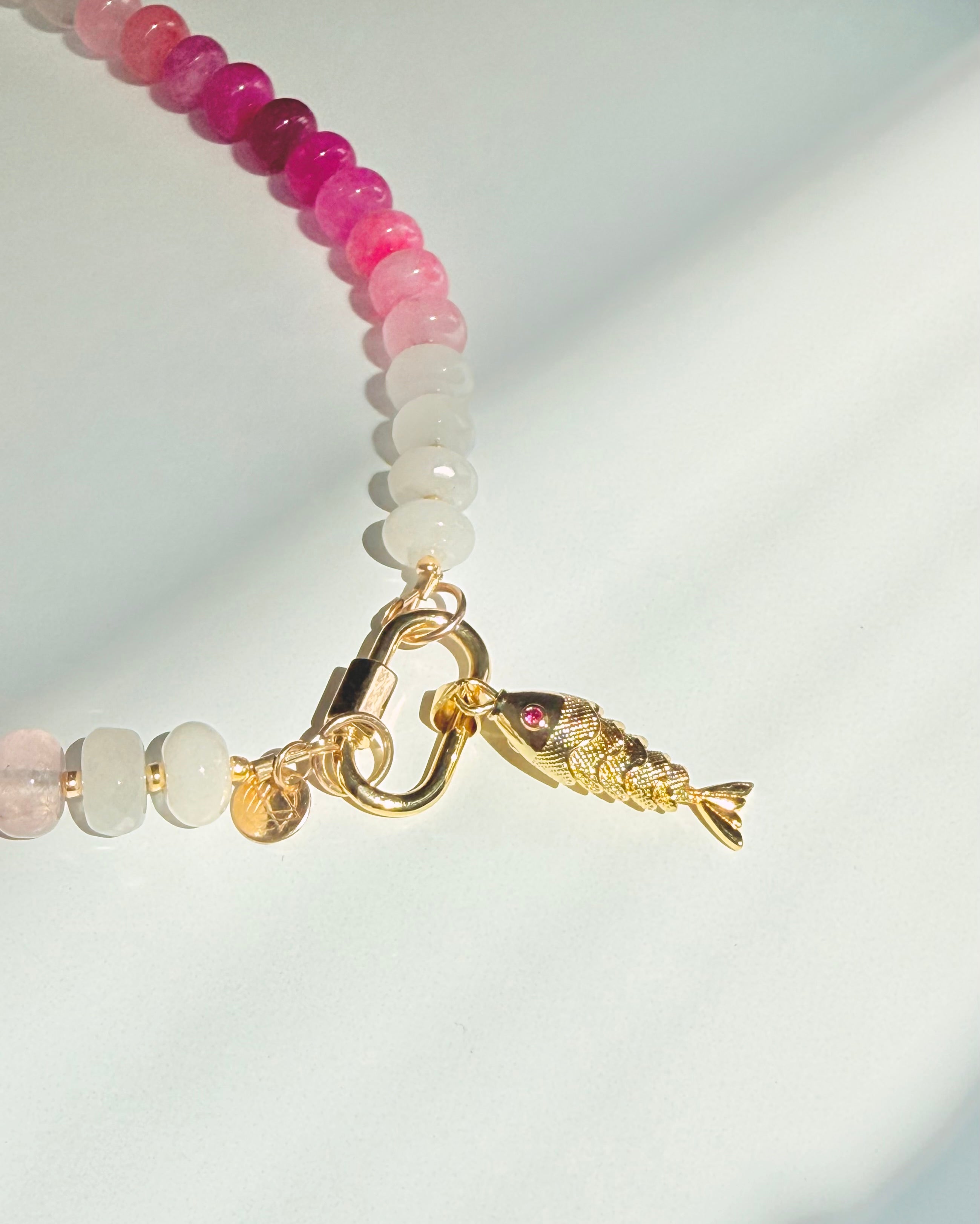 Close up of fish on Green pink and white stone beaded necklace with a gold carabiner and gold fish charm on a white background with a tropical leaf shadow