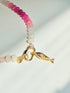 Close up of fish on Green pink and white stone beaded necklace with a gold carabiner and gold fish charm on a white background with a tropical leaf shadow