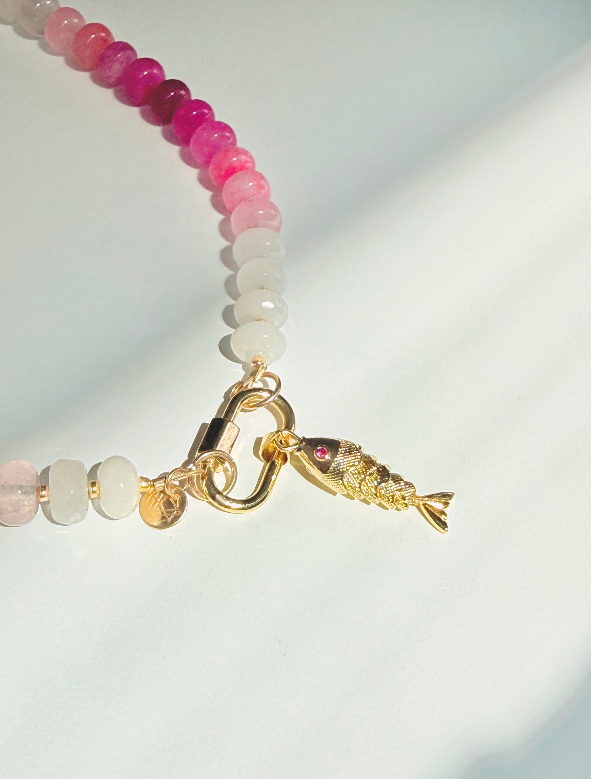 Close up of fish on Green pink and white stone beaded necklace with a gold carabiner and gold fish charm on a white background with a tropical leaf shadow